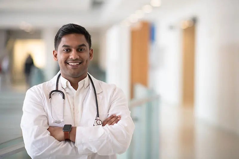 A male doctor stands in a hospital hallway smiling with his arms crossed