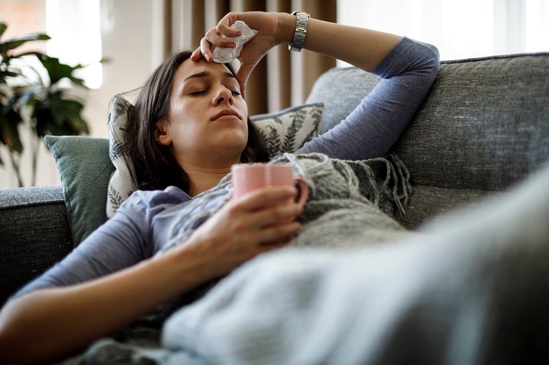 Woman sick on couch holding a mug and a tissue with her hand on her forehead.