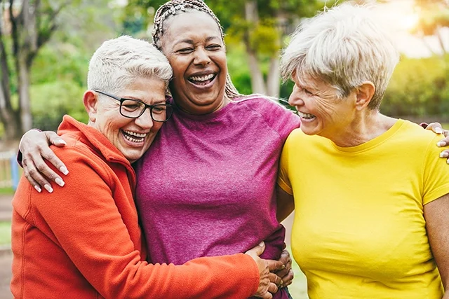 Three aged women embrace outside in a park. They're each wearing different-colored sweaters.