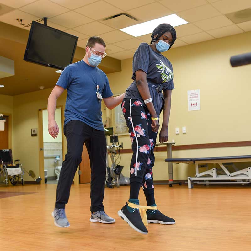 A rehab provider helps a woman learn to walk again. She's using bands around her ankles.