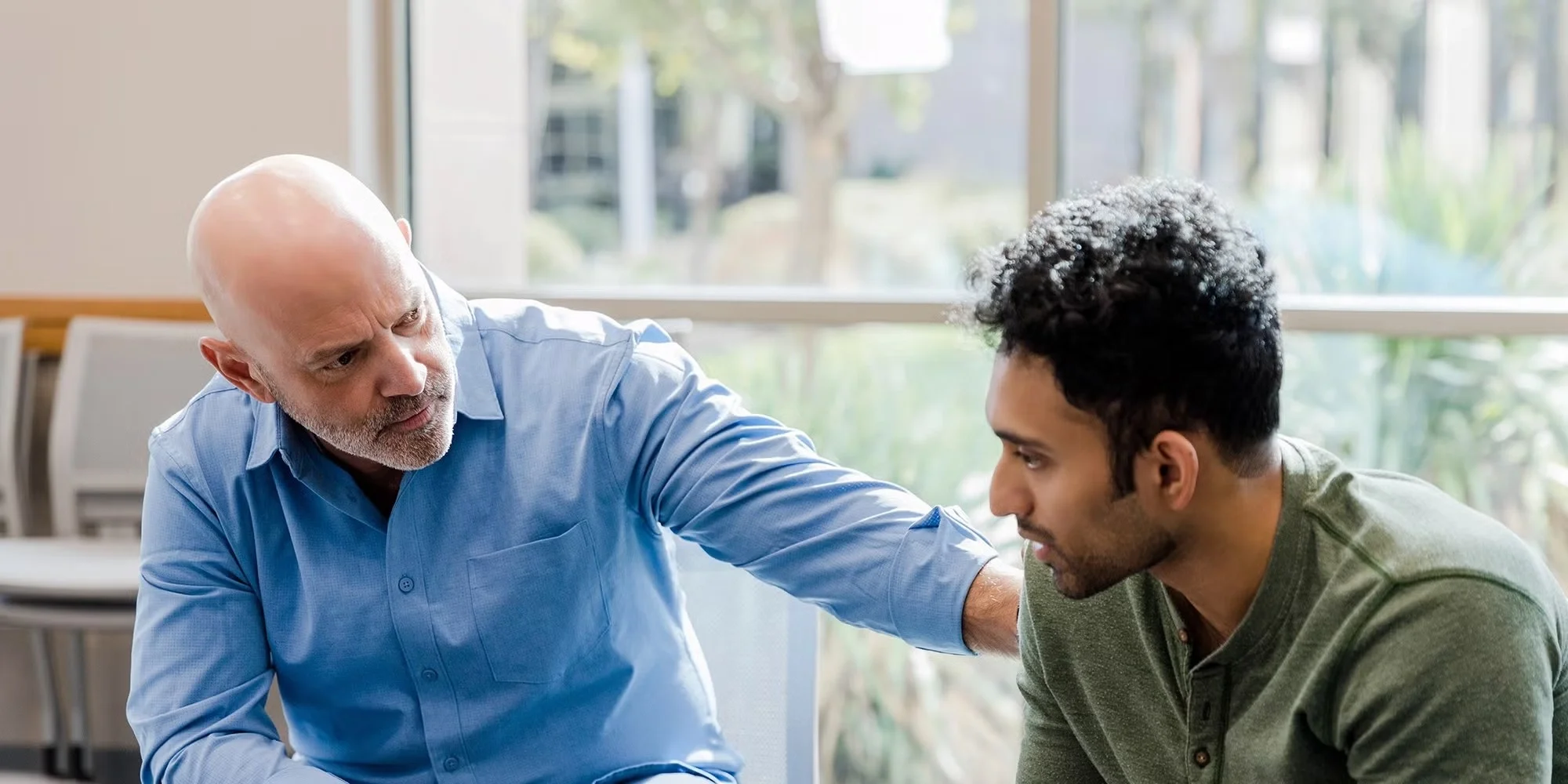 A man in a blue shirt gently consoles another man in a green shirt who appears to be down in the dumps