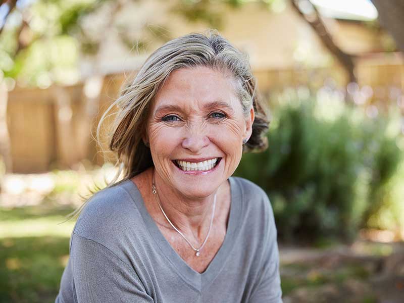 A senior woman appears happy and smiling in a backyard. It's bright outside with the sun shining. She's sitting in the shade.