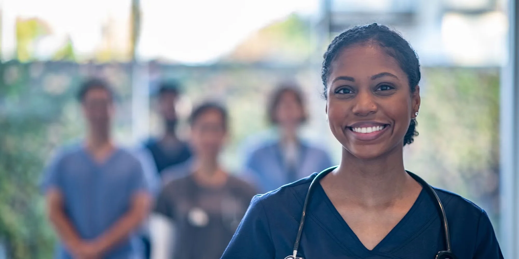 A group of medical residents stands in a hospital hallway. One woman in blue scrubs is closer to the camera, smiling happily.