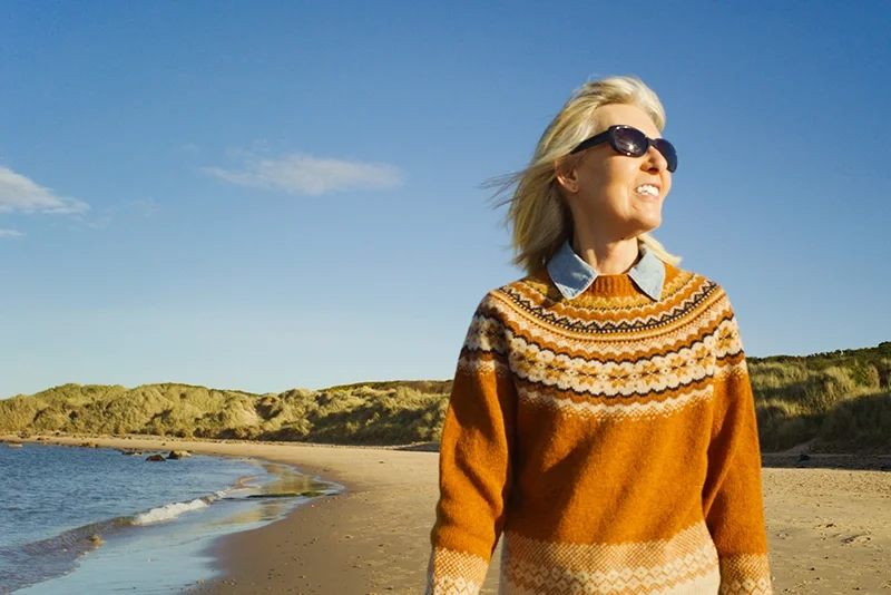 A woman gracefully scrolls along the beach in the fall. She's wearing a comfy orange sweater and sunglasses.