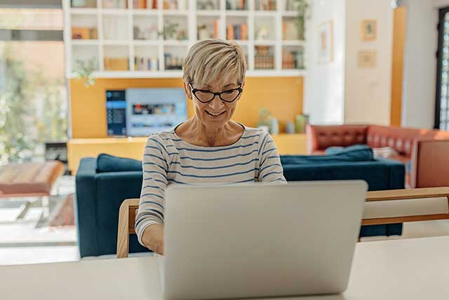 An older woman in a striped shirt sits at a laptop in her kitchen and schedules an appointment