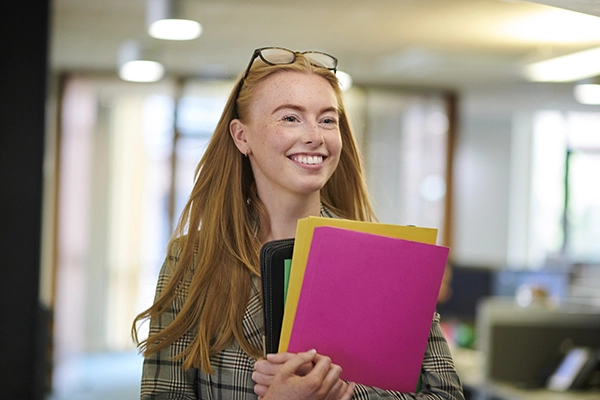 A young woman with red hair holds a bundle of file folders in a professional setting
