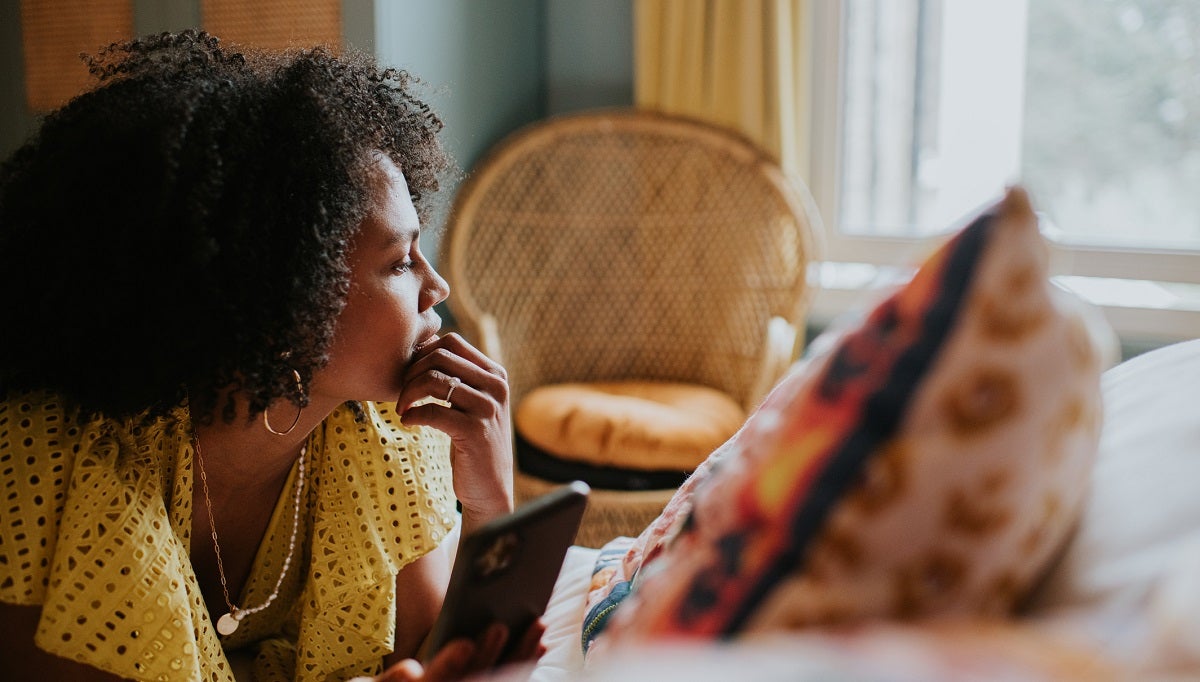 Woman sitting next to a window in yellow dress distracted from cell phone and deep in thought.