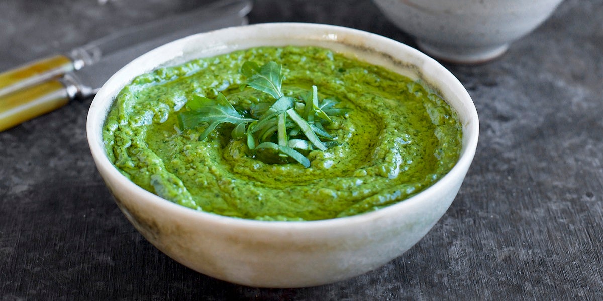 Green chive pesto in a white bowl on granite counter with butter knives and cloth napkins in background.
