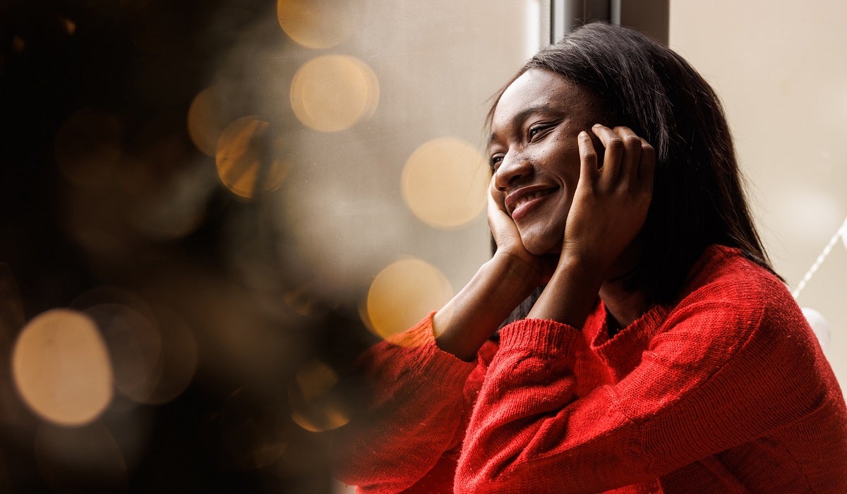 Young woman wearing red clothing, smiles while looking out the window with twinkling lights in the background.