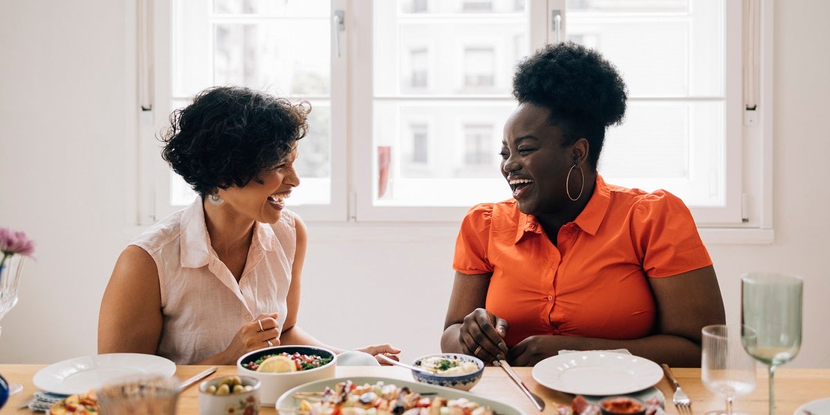 Two women laughing and having lively conversation while sharing a meal in a brightly lit room next to a window during the day