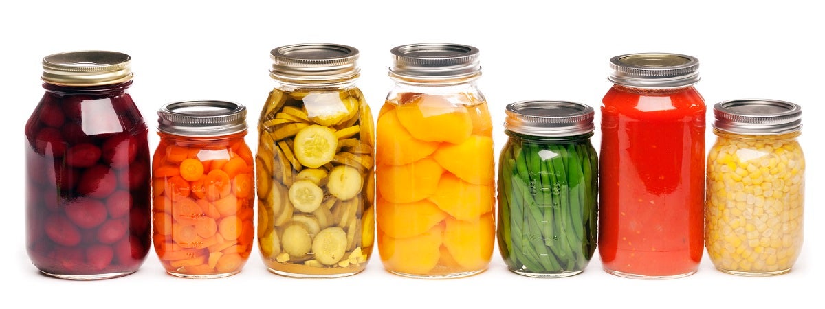 Seven glass jars that contain from left to right, beets, carrots, pickles, peaches, green beans, tomato sauce, and corn.