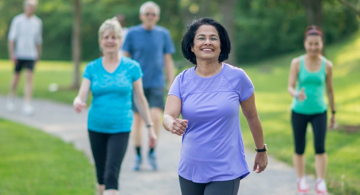 Mature woman wearing a purple exercise shirt walking outside with other people in a park.