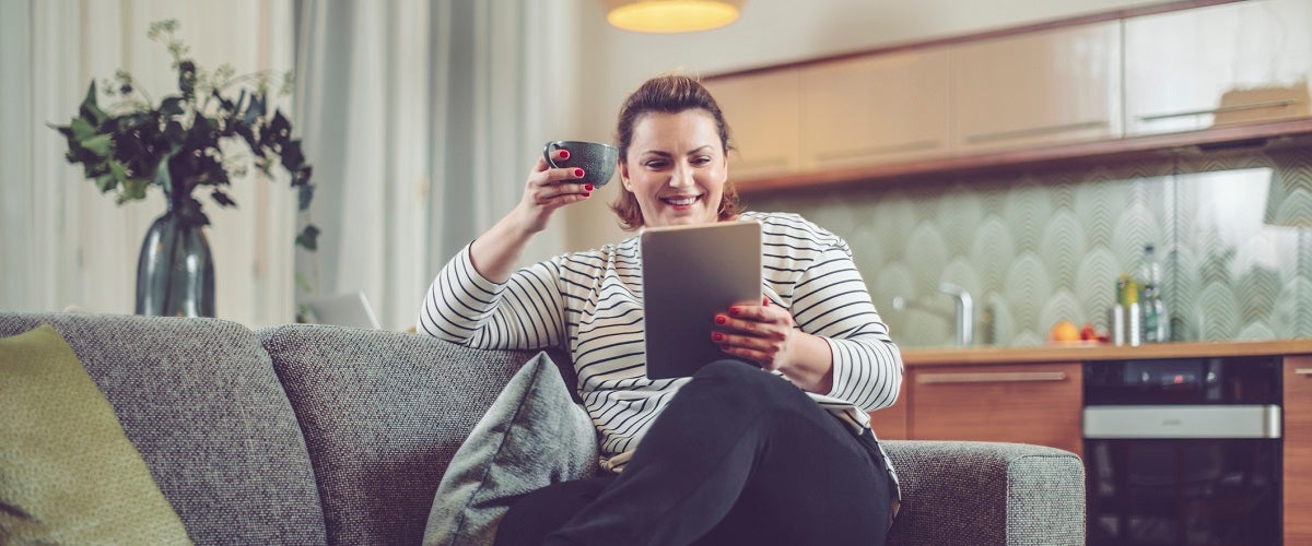 Woman sitting on couch reading a tablet and holding a mug.
