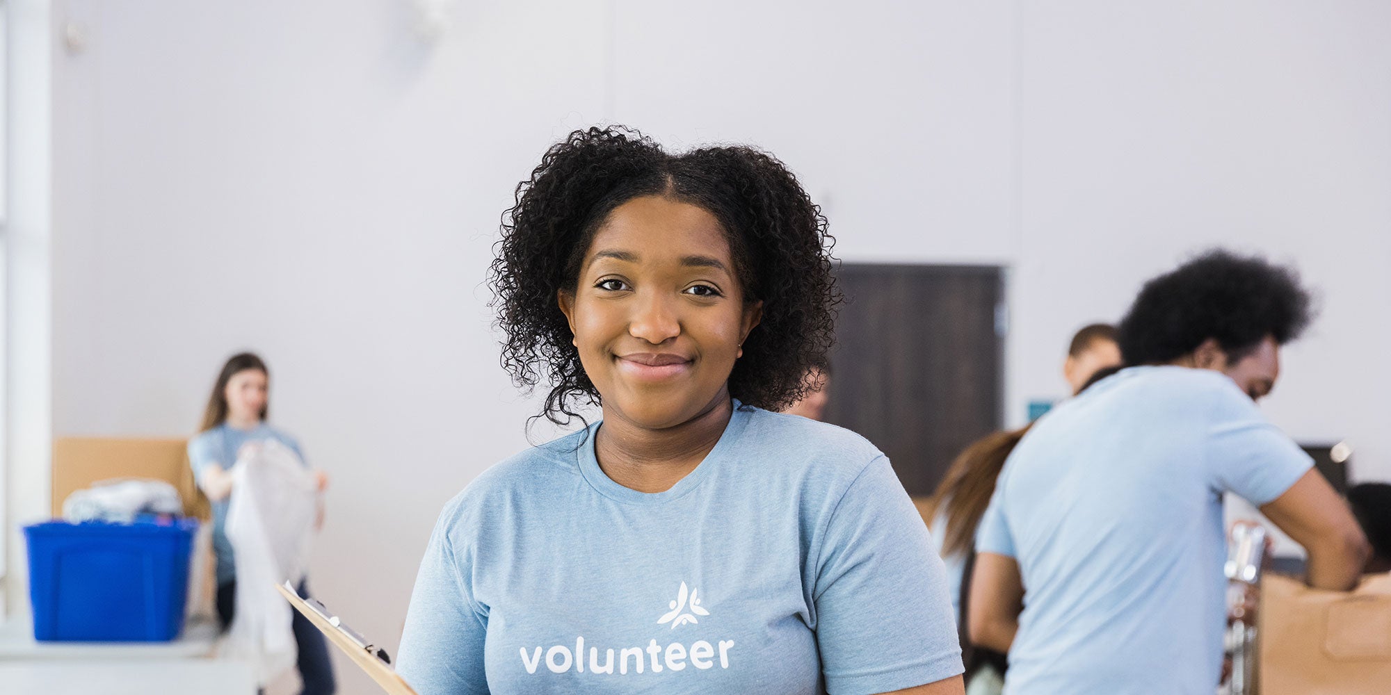 A young female volunteer stands with a clipboard in the middle of a room of other volunteers. They're collecting items in boxes.