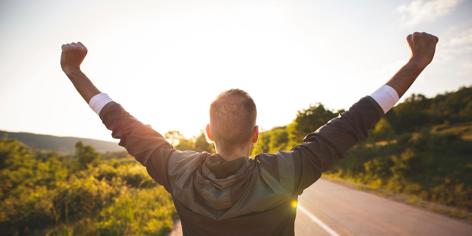 A man lifts his arms in celebration at the end of his run. He's in the middle of a street and the sun is beginning to set.