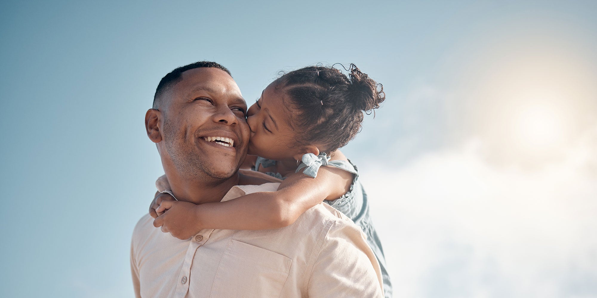 A young daughter rides on her father's back, kissing him on the cheek as he smiles lovingly
