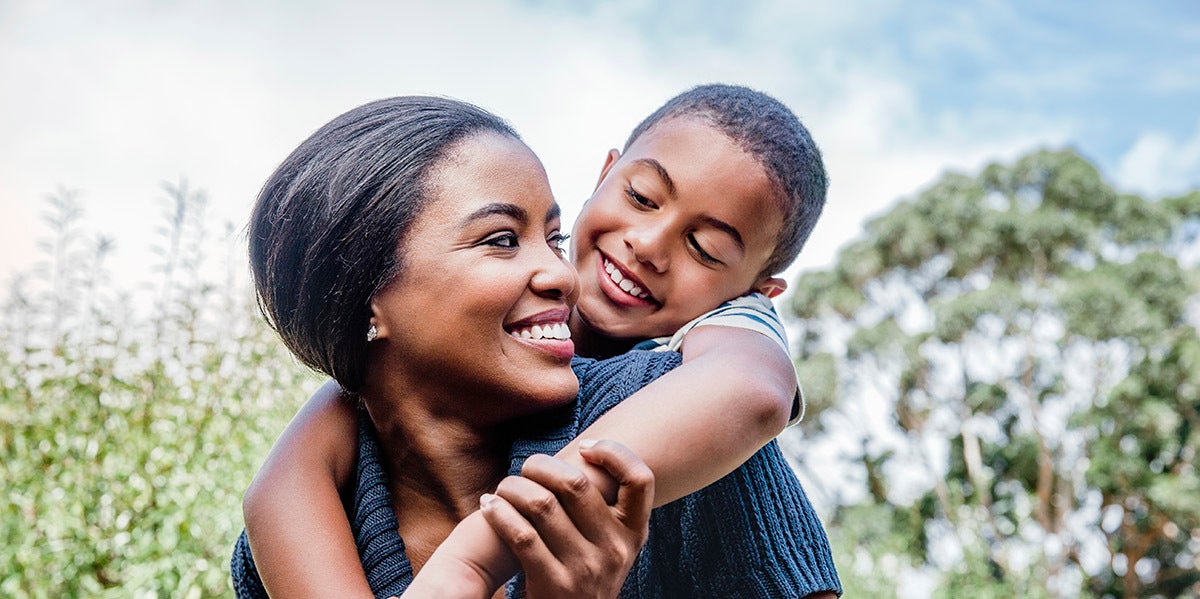A mother carries her son on her back, smiling together outside in a park