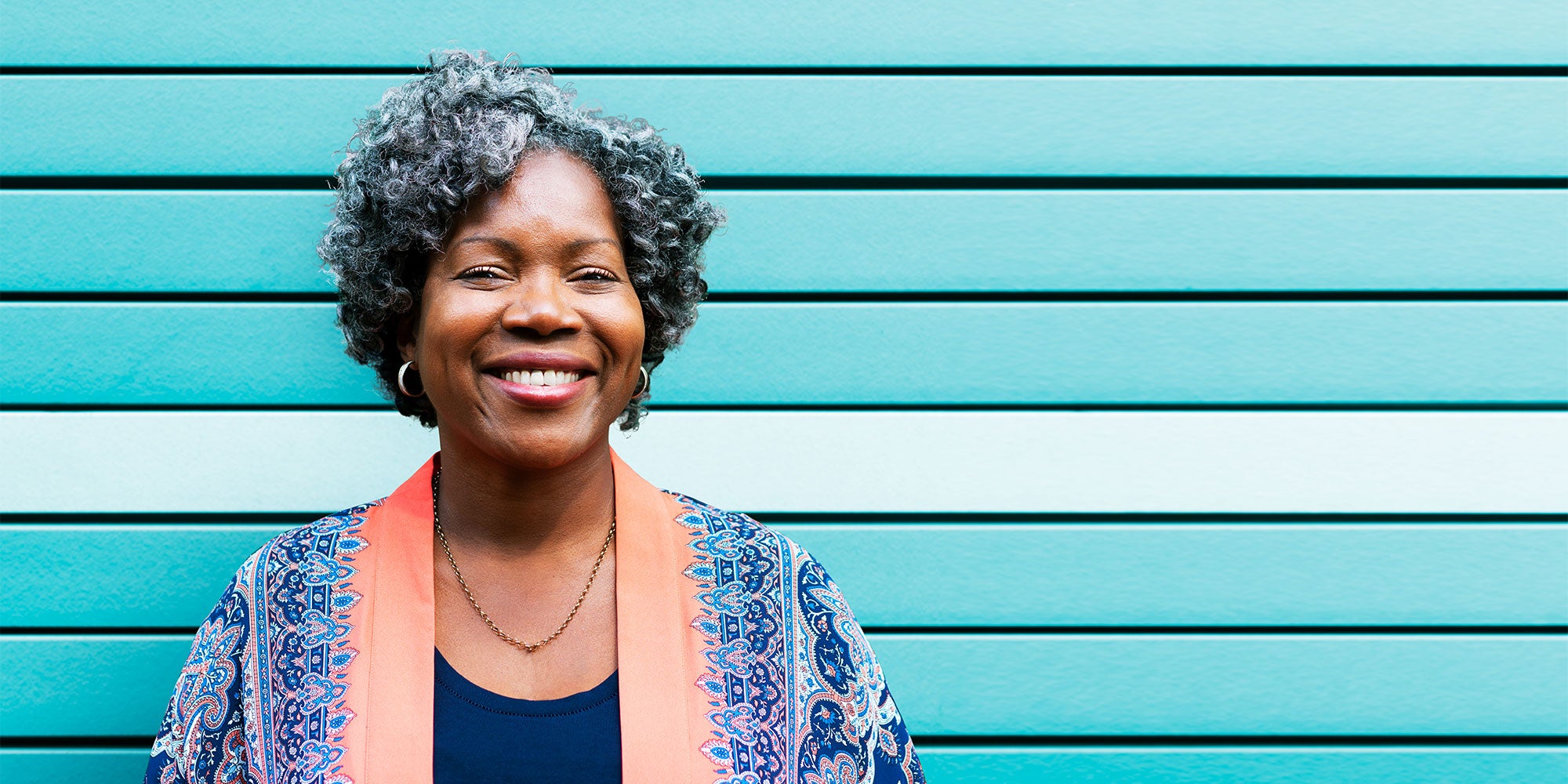 A woman stands in front of a light blue and white wall. She smiles, wearing gold hoop earrings and a pink and blue smock
