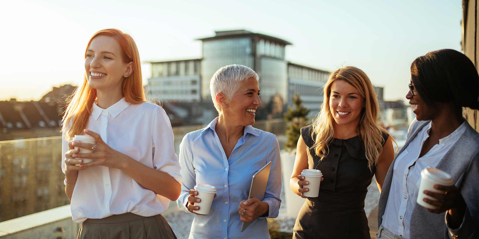 Four women chat as they walk along an exterior building walkway. They all also each have a cup of coffee.